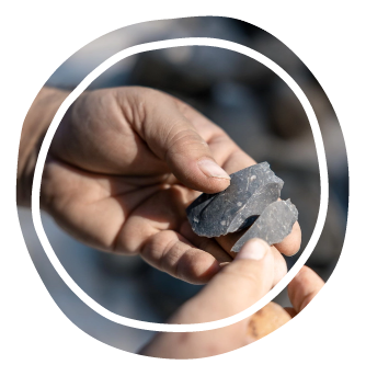 Two hands hold small dark rocks, shown against a blurred outdoor background.