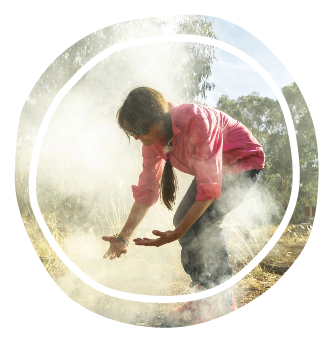 Woman in a pink shirt crouches outdoors, touching dusty ground as dust rises around her, within a circular white frame overlay.